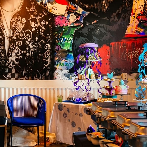 Colorful buffet setup with cupcakes, cakes, and chafing dishes against a vibrant mural.