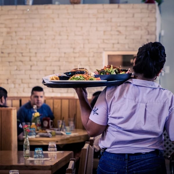 Waitress carries a tray with a burger and salad through a restaurant with brick walls.