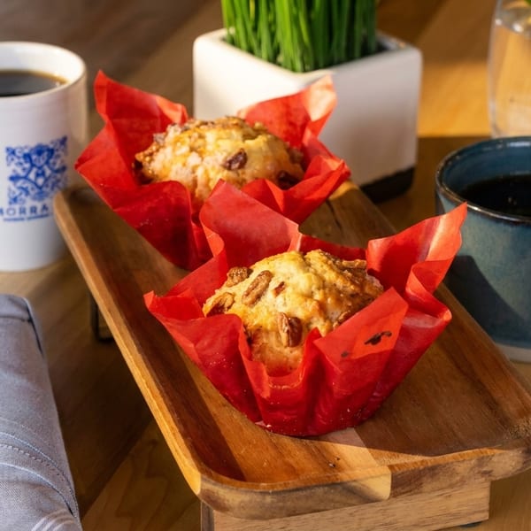 Two muffins in red wrappers on a wooden tray, with two cups of coffee and a plant.