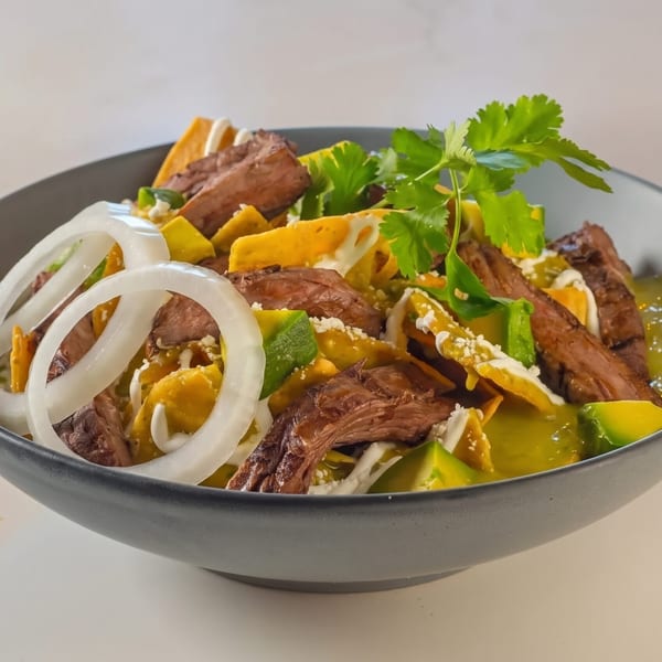 Chilaquiles with steak, avocado, onions, and cilantro in a dark bowl on a white surface.