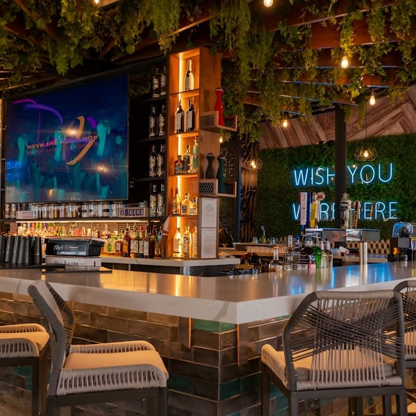 A vibrant bar with a large TV, liquor bottles, and a neon "WISH YOU WERE HERE" sign.