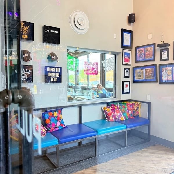 Restaurant interior with blue benches, colorful pillows, framed art, and a window looking into another dining area.