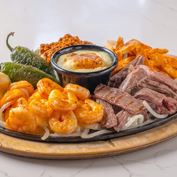 Sizzling fajita platter with shrimp, steak, onions, peppers, and a cheese dip on a wooden board.