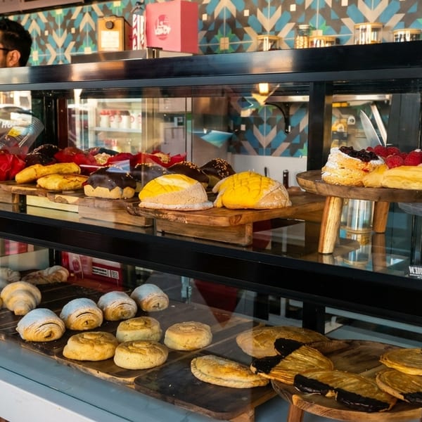 A bakery display case filled with various pastries, cakes, and breads, with a man in the background.