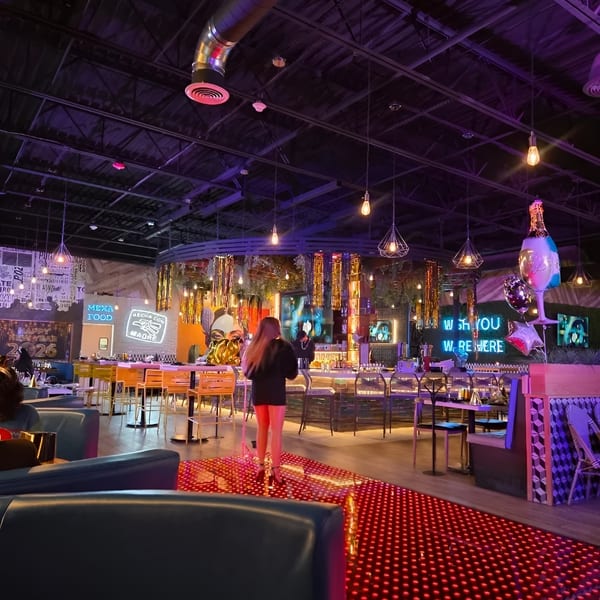 A woman walks on a red-lit floor in a vibrant, modern restaurant with neon signs and a bar.